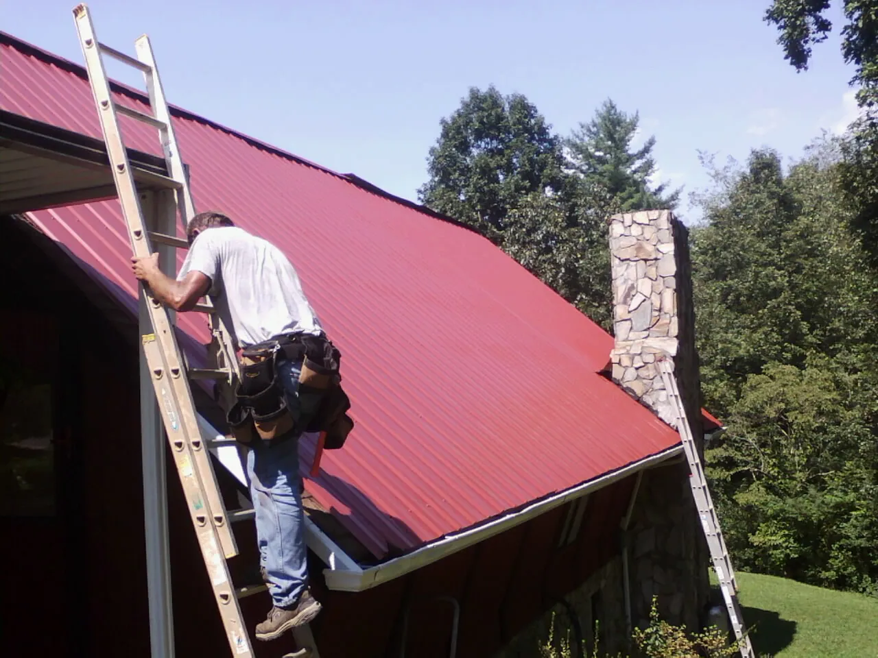 Roofer on a ladder beside a red metal roof and stone chimney — Soffit & Fascia Repair by Roofing Brothers in Glendo